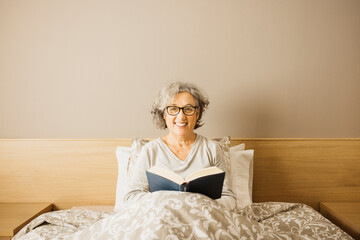 Portrait of a senior woman sitting on bed wearing pajama and reading a book in the morning.