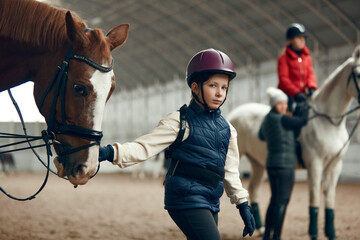 Little girl, kid in helmet holding horse and walking on special arena. Horseback riding course,...