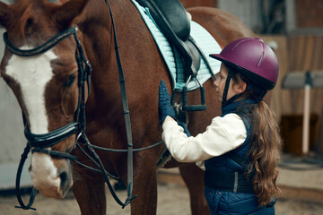 Little girl, kid in helmet taking care after horse, showing her love. Child training horseback riding in special arena, pavilion. Concept of sport, childhood, school, course, active lifestyle, hobby