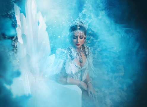Art photo real people Fantasy woman snow queen sits on ice throne chair, white long dress train bird feathers crown icicles. Fairy tale girl frozen princess creative costume. Winter lady nature fog