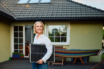 Sustainability concept. Portrait of happy mature woman standing outside her home with photovoltaic...
