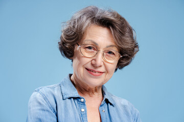Smiling 70 years old senior woman wearing eyeglasses and denim shirt isolated on blue background