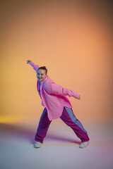 A young woman in a pink shirt poses against an orange studio background. The dancer demonstrates elements of jazz-funk choreography. Modern choreography. Full length.