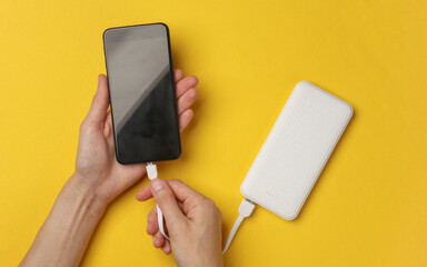Female hands connect a smartphone to an external battery power bank on a yellow background