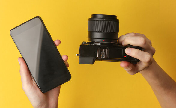 Hands holding a modern mirrorless camera and smartphone on yellow background