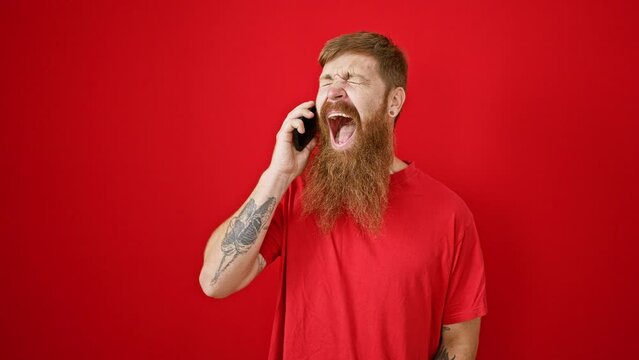 Overjoyed young redhead man ecstatically celebrating while speaking on the phone over an isolated red background