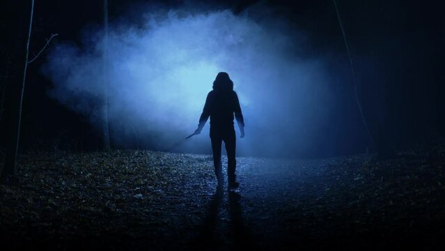 A terrifying maniac with a knife in his hand walks through an ominous forest at night. A black male figure emerges from the darkness.