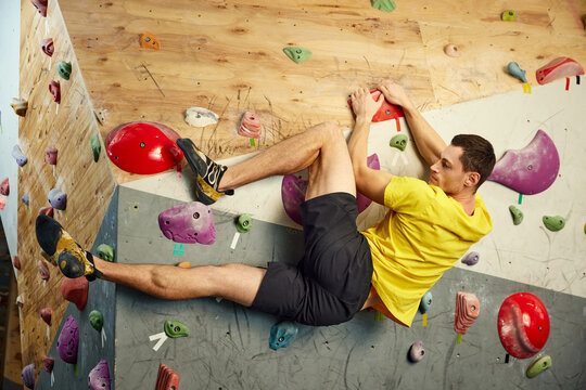 Muscular young man climbing artificial rocks on climbing wall, practicing bouldering activity indoors. Concept of bouldering, sport climbing, hobby, active lifestyle, school, training course