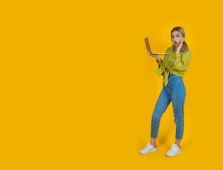 Shocked young woman, full body portrait of shocked young woman. Blonde girl hold use laptop amazed surprised isolated over studio yellow background copy space. Open mouth touching her face.