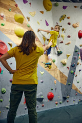 Fototapeta premium Female bouldering trainer teaching little girl, child bouldering activity, techniques. Little girl climbing wall, indoor class. Concept of sport climbing, hobby, active lifestyle, school, course