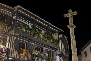 Fotografía nocturna de cruz de piedra y arquitectura tradicional con balcones de madera y tiestos...