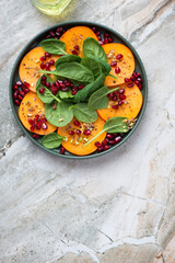 Fresh persimmon with baby spinach and pomegranate on a light-grey granite background, vertical shot with space, top view