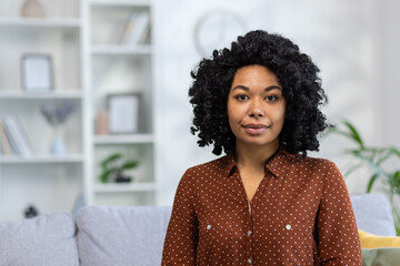 Close-up portrait of a serious young African-American woman sitting on the sofa at home and looking confidently at the camera - obrazy, fototapety, plakaty