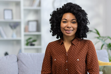 Portrait of a smiling young African American woman with curly hair sitting on the sofa at home and looking confidently at the camera. Close-up photo - obrazy, fototapety, plakaty