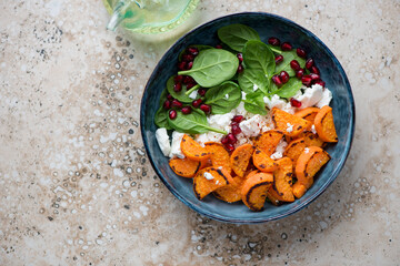 Blue bowl with roasted sweet potato, fresh spinach, feta and pomegranate, top view on a beige granite background, horizontal shot with space