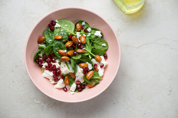 Roseate bowl with feta, baby spinach, pomegranate and almonds salad, above view on a light-beige stone background, horizontal shot