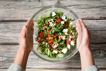 Female hands holding a glass bowl of vegetable salad with feta over a wooden background