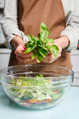 Female hands holding a bunch of green basil over a glass bowl with salad against the background of a kitchen