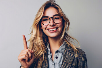 Cheerful young woman with glasses pointing upwards with one finger - obrazy, fototapety, plakaty