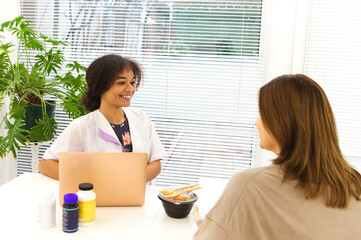 Smiling black lady doctor chatting with female patient at clinic advicing her vitamins