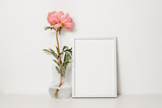 Picture frame mockup with peony flower in the vase on the white wooden table. Wooden white photo frame with copy space for design, cozy home interior