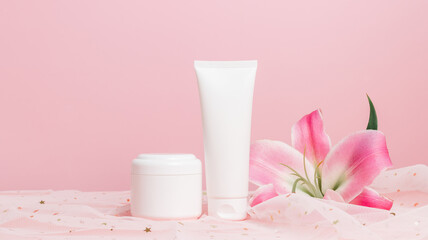 White tube of cream with cream bottle or lotion on glittering textile with pink lily flowers, copy space, banner size. Cream containers mockup, skincare, pink gentle background