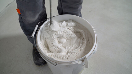 White solution in a bucket, mixed with a mixer. A worker mixes plaster in a bucket to level and putty the walls of an apartment using an electric drill, close-up. Mixing white plaster in a bucket with
