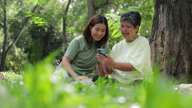 An elderly mother sits and relaxes with her daughter in the garden, looking at photos on a smartphone together. Caring for the elderly to be happy in retirement. Happy time. Asian family concept