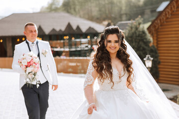 A beautiful young bride, in a summer park, walks ahead of her groom. Beautiful wedding white dress. Walks in the park. A happy and loving couple.