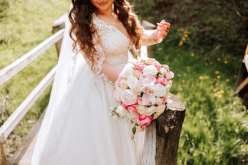Obraz na płótnie Canvas Curly brunette bride poses with a bouquet, near a wooden railing. Magnificent dress with long sleeves, open bust. Spring wedding