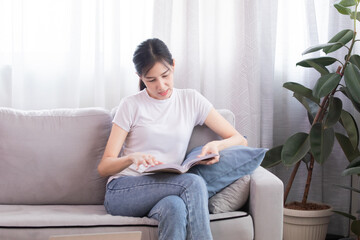 Asian single mother female reading favorite literature book sitting on sofa relaxing on holiday weekend, freelance creative design work from home enjoy free time leisure indoors in cozy living room