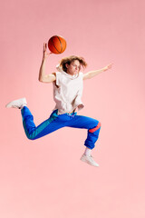 Young man dressed in retro sportwear sitting and posing with basketball ball against pastel pink studio background. Concept of active lifestyle, sport and recreation, hobby, goal, win.