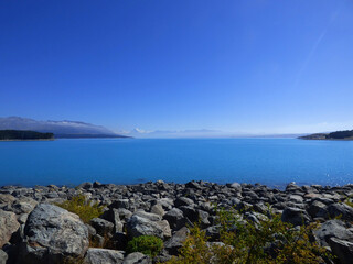 View over lake Tekapo with Southern Alps and Mt. Cook, heighest mountain of New Zealand, in the background. Rocky beach and clear blue water on summer day.