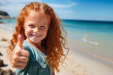 Happy red-haired child girl showing thumb up on the beach. Summer holidays concept.