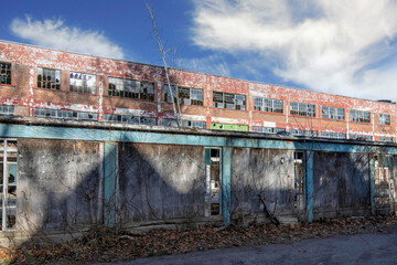 Facade of a derelict former factory building with broken windows, broken wooden fence in foreground, blue  sky, nobody	