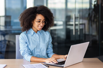 A young African-American woman is sitting in the office at a desk, working and typing on a laptop.