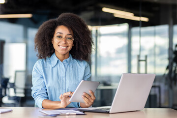 Portrait of a smiling young African American woman sitting at a desk in the office, holding a tablet and smiling at the camera.