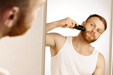 Man in T-shirt in bathroom stands in front of mirror for facial grooming, wellness and skincare treatments at home. Concept of natural beauty, selfcare treatment, male facial care, wellness. Ad - Powered by Adobe