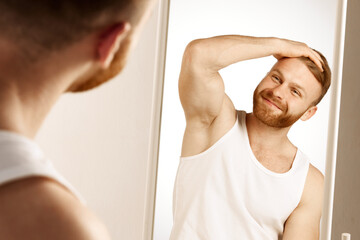 Attractive red haired, bearded man touching face applying moisturizer on face, standing near mirror...