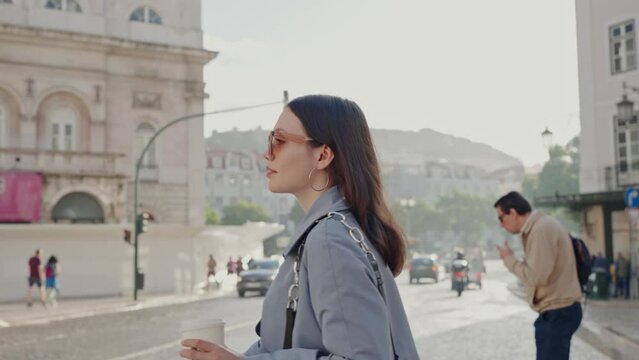 Young woman in stylish outfit and trendy sunglasses go to work and crossing road on pedestrian walkway. Business lady walk on street in city with cup of coffee.
