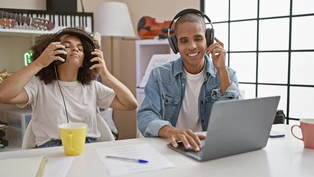 Two confident musicians joyfully listening to their own song using a laptop in a cozy radio studio, radiating happiness and raw talent