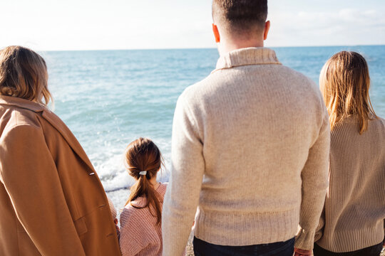 Happy family against backdrop of sea. Mom, dad, teen and preschool girl standing back. People are dressed in warm sweaters and coats. Man, woman, teennager and little child look at ocean water.