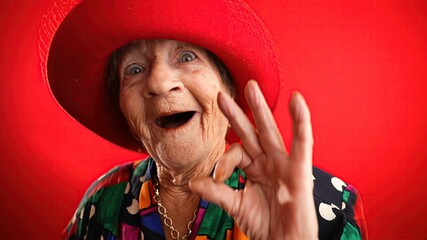 Happy fisheye portrait caricature of funny elderly woman with red hat giving OK sign gesture with...