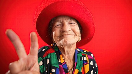 Portrait of funny, elderly mature woman, 80s, giving peace sign gesture with no teeth and red hat, isolated on purple background studio. Concept of youthful old lady with copy space