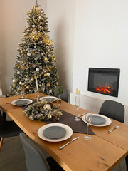 A festive dining room with a snow-frosted Christmas tree decorated with gold and white ornaments, a set dining table with elegant gray plates and glassware, and a lit fireplace in the background. 