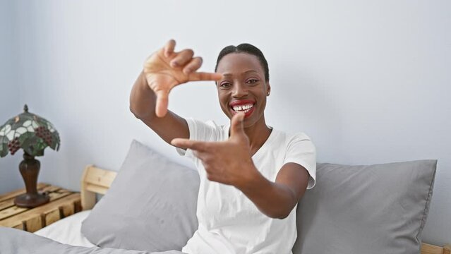 Happy african american woman in bedroom, lying on bed, beautifully framing her smiling face with creative hand gesture, conveying a concept of photography