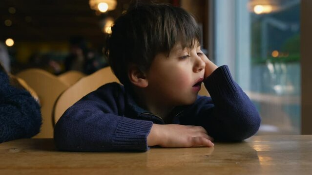 Bored small boy yawning while waiting at restaurant table by window. Child feeling boredom in authentic lifestyle moment awaiting for food to arrive
