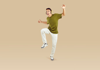 Fototapeta na wymiar Full length portrait of a young joyful excited happy boy student wearing casual clothes having fun, dancing and listening to music in headphones isolated on a studio beige background.