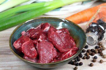 Raw deer meat for venison ragout or goulash. Bowl with pieces of deer meat, fresh vegetable,  juniper berries and allspice for cooking around.