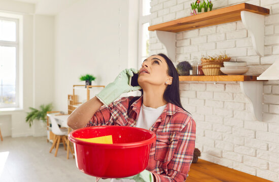Having a roof leak at home. Worried young woman in rubber gloves holding red plastic basin, looking up at water dripping down from ruined ceiling and talking to repair and maintenance service on phone